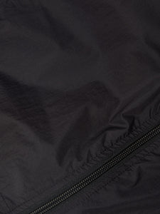 Women's ultra-lightweight running jacket (55g/m²) in Black. It features an adjustable hood, two side pockets, a back pocket, and side vents for enhanced ventilation. It is equipped with adjustable straps at the hem for a customizable fit. Made from 100% recycled nylon, it offers protection against wind and light rain. Additionally, it is packable, PFC-free, and features a reflective UNNA logo on the back, along with a UNNA patch on the zipper